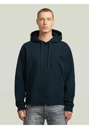 Jeune homme aux cheveux courts, portant un sweat à capuche bleu marine foncé et un jean gris clair usé, debout devant un fond clair uni.