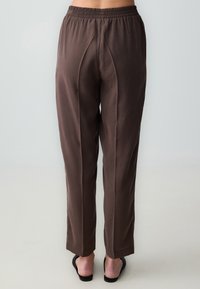 Bruine, lichte broeken met een elastische tailleband, rechte pasvorm en voorplooien; de stof heeft een gladde textuur.