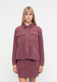 Σακάκι από κορδούρα σε σκόνη ροζ χρώματος με γιακά, κουμπιά μπροστά και δύο τσέπες στο στήθος, σε συνδυασμό με μια ταιριαστή υφασμάτινη φούστα.