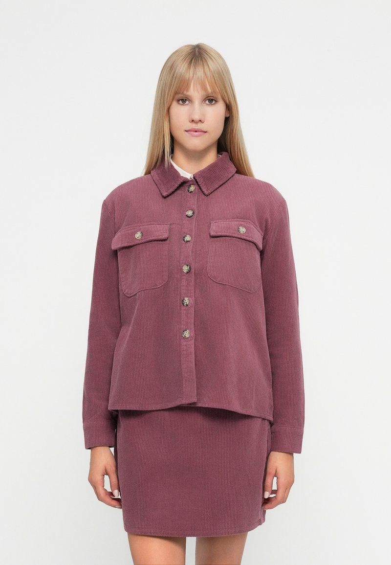 Σακάκι από κορδούρα σε σκόνη ροζ χρώματος με γιακά, κουμπιά μπροστά και δύο τσέπες στο στήθος, σε συνδυασμό με μια ταιριαστή υφασμάτινη φούστα.