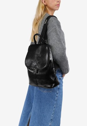 Mochila de cuero negro con solapa redondeada, cierre de cremallera y asa superior. Textura suave con detalles de costura visibles.