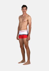 Shorts de bain rouges et blancs avec un cordon de serrage à la taille, présentant une bande noire accentuée à l'horizontale. Matière lisse, design ajusté.