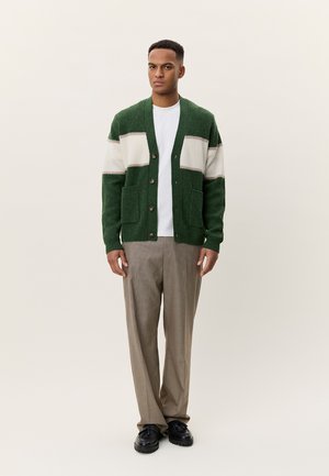 Green and cream striped cardigan with textured fabric, featuring two front pockets and brown buttons, worn over a white shirt and beige trousers.
