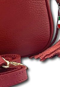 Eck einer roten Lederhandtasche mit strukturiertem Oberflächenmaterial, passendem Riemen mit goldenen Verschluss und einem Quasten-Schlüsselanhänger in den Farben der italienischen Flagge.