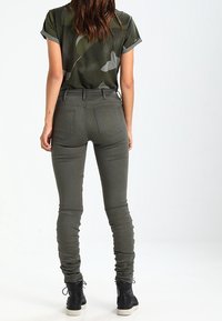 Olivgrön tajta jeans med en slät textur, fem fickor och bälten, tillsammans med en kortärmad mönstrad topp. Svarta ankelstövlar.