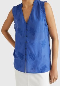 Haut sans manches bleu avec un décolleté en V profond, bords de épaules volants et broderies florales. Il présente une fermeture à boutons et une texture douce.