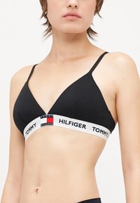Bralette triangulaire en coton noir avec des fines bretelles, présentant un bandeau élastique de marque blanc avec des accents rouges, blancs et bleus. Texture lisse.