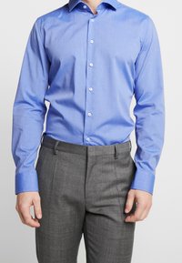 Světle modrá košile s dlouhým rukávem a zapínáním na knoflíky, kontrastní manžety a šedé kalhoty s upraveným střihem a strukturovaným povrchem.
