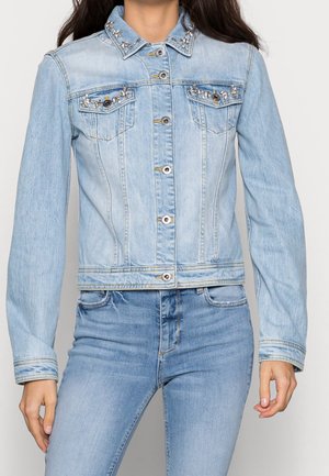 Kvinde iført lyseblå denimjakke med rhinstenudsmykning på krave og brystlommer, stylet med matchende denimjeans.