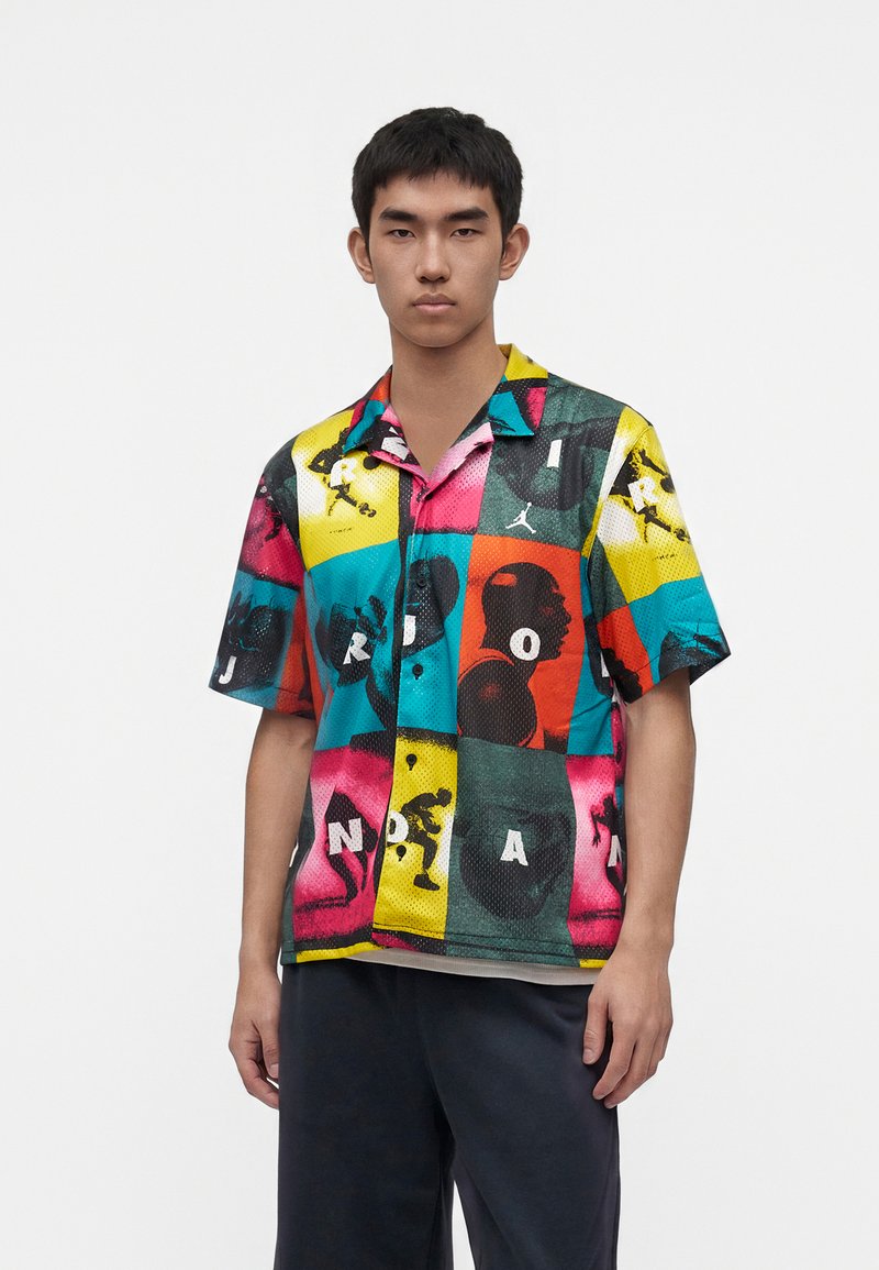 Jeune homme portant une chemise colorée à manches courtes avec une silhouette multicolore de basket-ball et des motifs de lettres, debout devant un fond uni.