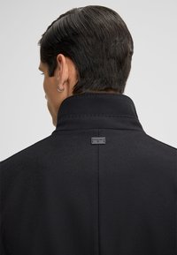 Mann mit kurzen dunklen Haaren und einem silbernen Creolenohrring, der einen schwarzen Hochkragenmantel trägt und von hinten vor einem schlichten Hintergrund zu sehen ist.