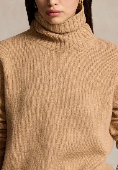 Nő, aki bézs bordás garbót visel arany karika fülbevalókkal, nyaktól derékig látható semleges háttér előtt.