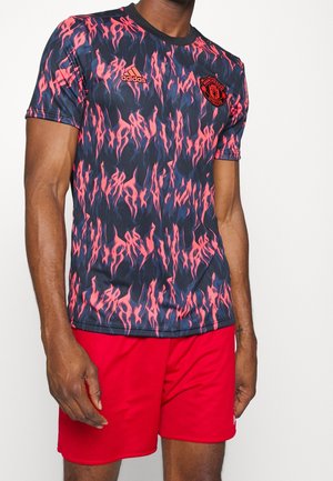 Mann trägt ein schwarzes und pinkes Manchester United Adidas Shirt mit Flammenmuster und roten Shorts vor einfarbigem Hintergrund.