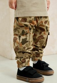 Pantaloni cargo con motivo mimetico in tonalità terra, dotati di polsini elasticizzati e tasche laterali, abbinati a scarpe da ginnastica nere e calze bianche.