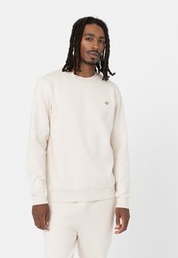 Crème-kleurige sweatshirt gemaakt van zachte stof met een geribbelde kraag en manchetten, voorzien van een klein, kleurrijk logo op de borst.