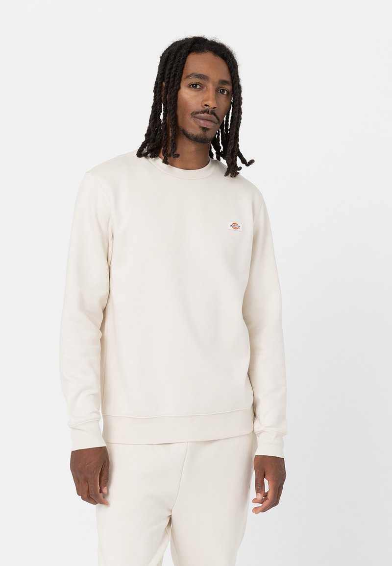 Crème-kleurige sweatshirt gemaakt van zachte stof met een geribbelde kraag en manchetten, voorzien van een klein, kleurrijk logo op de borst.