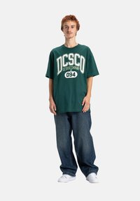Zielona bawełniana koszulka z białym napisem "DC Shoe Company 094" z przodu, zestawiona z luźnymi niebieskimi dżinsami i białymi sneakersami.