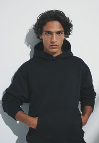 Tmavomodrá mikina s kapucňou z bavlnenej zmesi, s vreckom na bruško, rebrovanými manžetami a šnúrkou na kapucni, ktorú nosí modelka na bielom pozadí.