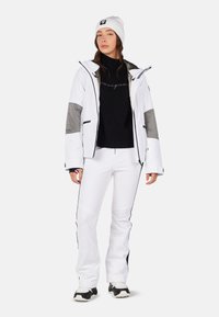 Giacca da sci bianca con dettagli grigi, maglione nero lavorato a maglia, pantaloni bianchi con piping nero e scarpe sportive nere. Berretto con logo.