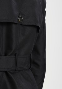 Black trench coat featuring a buttoned flap, textured fabric, and a matching belt. The design emphasizes a tailored fit and detailed stitching.