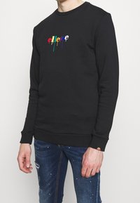 Felpa nera con logo colorato che cola. Presenta un collo rotondo, maniche lunghe e polsini a costine. Indossata con jeans in denim strappati.