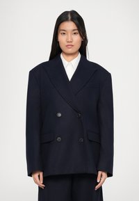 Frau mit langen schwarzen Haaren, die einen übergroßen, dunkelblauen, doppelt knöpfbaren Blazer über einem weißen Hemd mit Kragen trägt und vor einem schlichten Hintergrund steht.