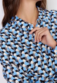 Camisa abotonada con un patrón geométrico en azul, negro, crema y gris. La tela parece suave con un cuello y puños con solapa.