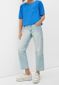 Blaues kurzärmliges Oberteil mit einer Fronttasche, kombiniert mit hellblauen geraden Jeans und weißen Sneakers mit strukturierten Akzenten.