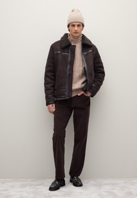 Braune Lammfelljacke mit schwarzen Lederakzenten, beiger Strickpullover, braune Cordhosen und schwarze Schuhe. Modell posiert vor neutralem Hintergrund.