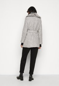 Abrigo gris texturizado con cuello de piel sintética, cintura ceñida con cinturón y mangas largas, combinado con pantalones negros remangados y botas de tobillo.