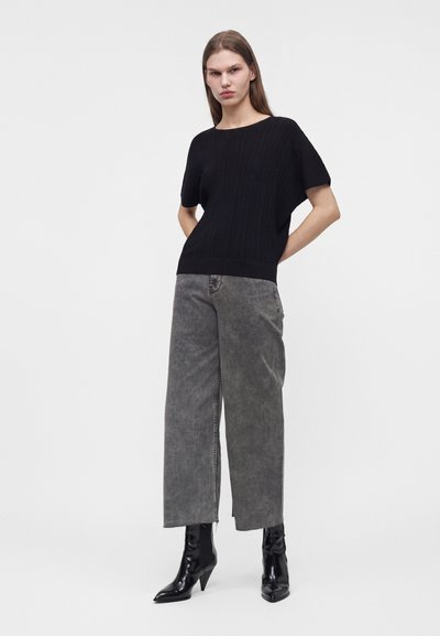 Femme debout portant un haut noir à manches courtes, un jean gris large et court, et des bottines noires à talons pointus.