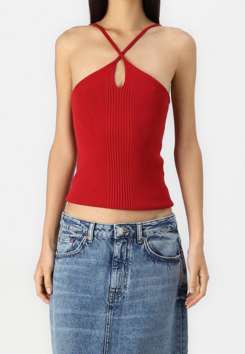 Femme portant un haut dos nu en maille côtelée rouge avec des bretelles croisées et une ouverture en forme de trou de serrure à l’avant, associée à une jupe en jean bleue.