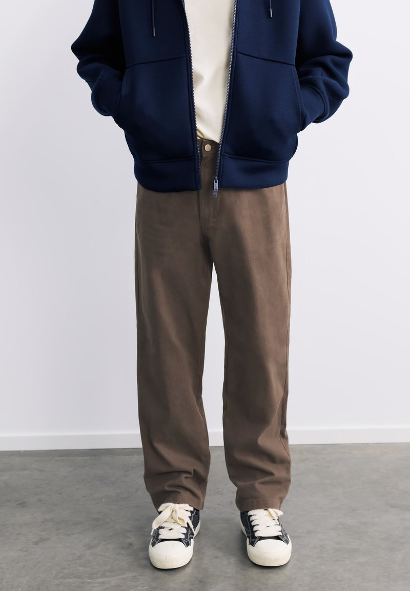 Personne portant un sweat à capuche bleu marine zippé, une chemise beige, un pantalon marron et des baskets noires et blanches, debout sur un sol gris contre un mur blanc.