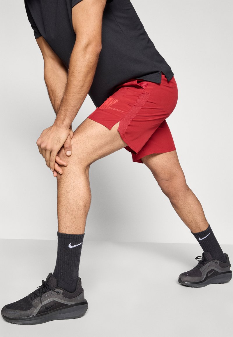 Pantaloncini atletici rossi con spacchi laterali, abbinati a calzini sportivi neri e scarpe da ginnastica nere. Caratteristiche includono tessuto testurizzato e dettagli con logo.