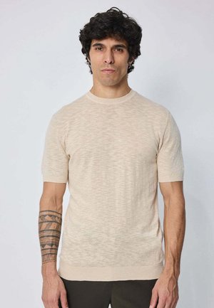 Homme aux cheveux bouclés noirs portant un pull beige à manches courtes et un pantalon noir, montrant un tatouage géométrique noir sur l'avant-bras gauche, debout contre un mur blanc.