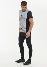 Mans atletiska outfit som består av en ärmlös, silverreflekterande väst och en svart kortärmad tröja, tillsammans med svarta leggings och cykelskor.