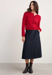 Sarkans adīts džemperis ar apaļu izgriezumu un pogu aizdari, apvienots ar garu tumšo denima svārku ar vertikālām šuvēm un kabatām. Brūni potīšu zābaki.