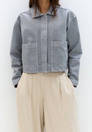 Woman wearing a light gray cropped jacket with front pockets and loose beige high-waist pants, hands in pockets, against plain background.