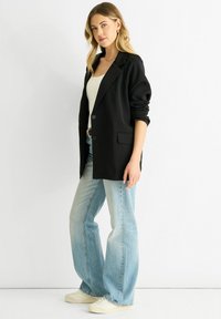 Black tailored blazer with shoulder padding, single button closure, and side pockets, paired with light blue flared jeans and white sneakers.