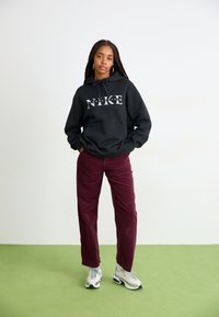 Crna dukserica s "NIKE" logotipom, uparen s burdunim kord ili hlačama i sivimtenisicama. Model ima dugu pletenicu i naušnice na obruču.