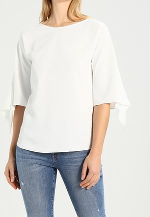 Femme portant un chemisier blanc à manches cloches 3/4 et un jean bleu clair, debout devant un fond clair uni.
