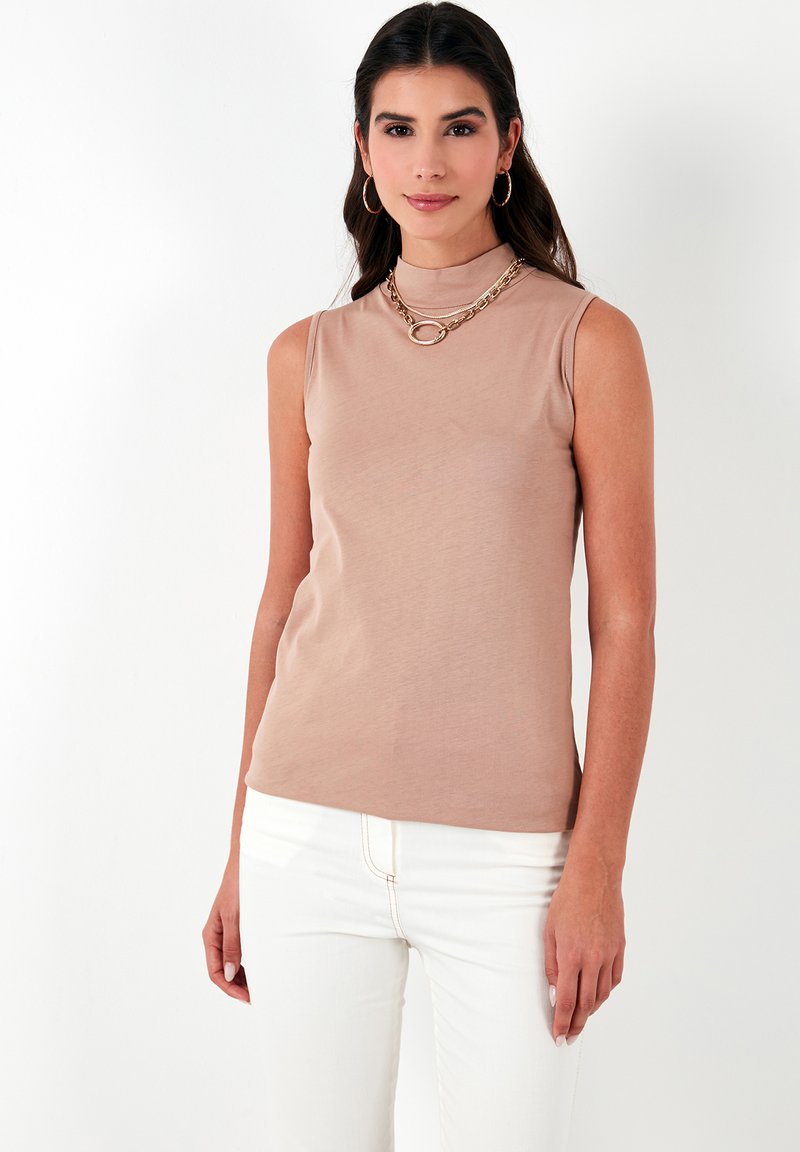 Sleeveless, fitted top in light beige, made of smooth fabric. Features a mock neck and seamless design. Paired with white jeans.