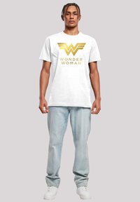 Balta kokvilnas T-kreklu ar zeltītu Wonder Woman logo un tekstu. Modelis valkā gaiši zilo džinsus un baltas sporta kedas.