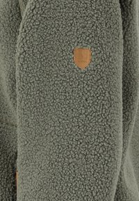 Fleece-takki oliivinvihreänä, jossa on teksturoitu pinta ja nahkabrändilogo. Vetoketj yksityiskohtia sivusaumassa.