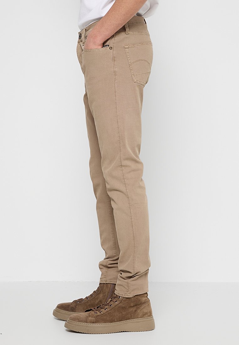 Beżowe spodnie slim fit o gładkiej fakturze, z pięcioma kieszeniami i subtelnym detalem szwu. Zestawione z brązowymi zamszowymi butami za kostkę.
