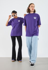 Camisetas de algodón moradas con un pequeño gráfico en el lado izquierdo del pecho, combinadas con pantalones acampanados negros y jeans de color azul claro. Gafas de sol blancas y zapatos casuales.
