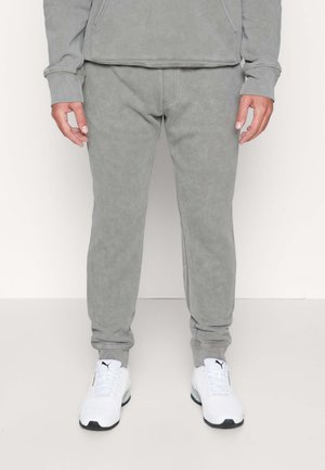 Persona che indossa pantaloni da jogging grigi e sneakers bianche Puma, in piedi davanti a uno sfondo chiaro e semplice.