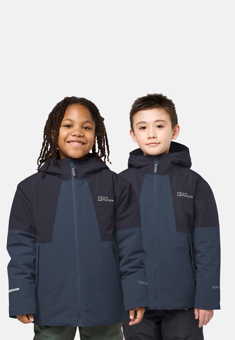 Zwei Kinder tragen dunkelblaue Jack Wolfskin Jacken mit Kapuzen und stehen nebeneinander, eines lächelt, das andere hat einen neutralen Gesichtsausdruck.