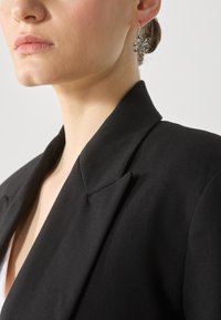 Blazer negru cu o guler ascuțit, purtat peste un top alb. Prezintă un cercei cu model floral. Textură de țesătură netedă și croială ajustată.