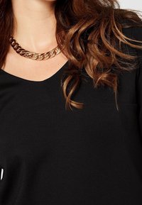 Svart v-ringad topp med korta ärmar, med en liten ficka. Framhävd av ett tjockt guldkedjehalsband och texturerat, vågigt hår.